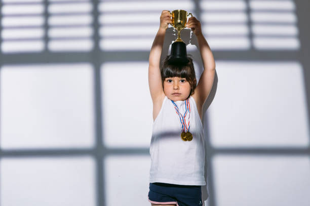 ragazza dai capelli scuri con medaglie del campionato sportivo, alzando la sua coppa dei trofei con le mani, sopra le ombre di una finestra con l'otturatore sopra di lei. concetto di sport e vittoria - humor athlete trophy one person foto e immagini stock