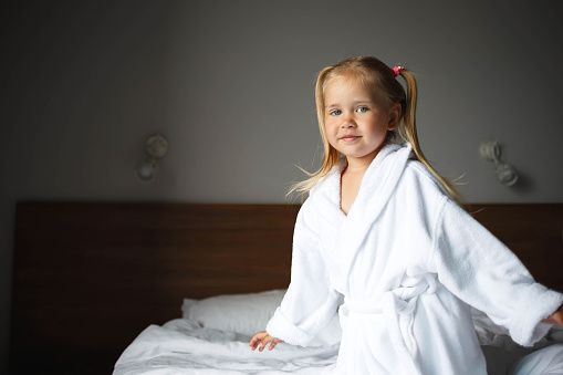 portrait of a little cute girl in a white robe lying on the bed in the bedroom.