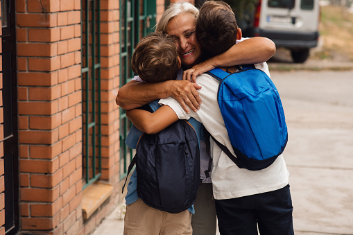 Mature adult blond lady is kissing and hugging her grandkids. Two boys are ready to go to school, and granny is saying goodbye to them wishing them good luck. They are smiling, feeling good because of first school day. Rear view.