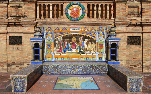Image with the name of the spanish city of Pamplona with a historical scene painted on ceramic tiles, with and the emblem shield above - seating benches in Spain Square in Seville