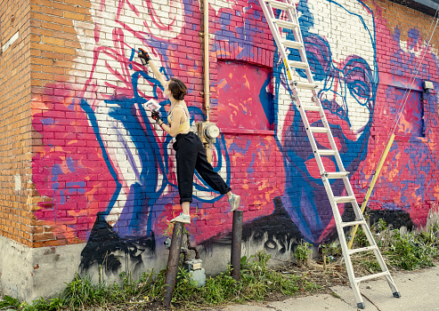 Young woman painting mural using spray paints. She is dressed in casual work clothes. Exterior of urban street, back alley with old brick wall of the house.