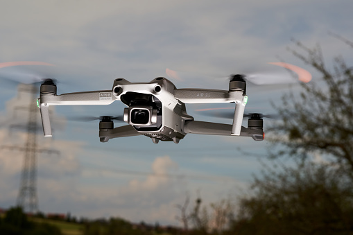 Nürtingen, Germany - June 26, 2021: Drone air 2s from the dji company hovers in the air. Electricity pylon and bare tree in front of a blue cloudy sky. Low angle view.