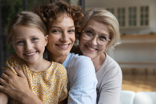 Happy emotional affectionate young woman posing with cute preschool child daughter and loving older mature mother at home, joyful sincere different female generations family showing tender feelings.