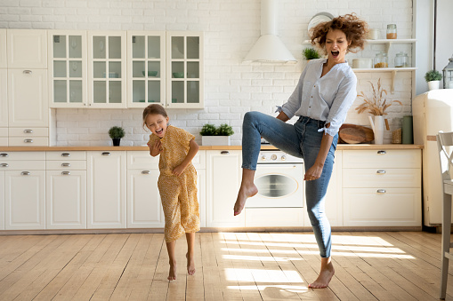 Full length overjoyed crazy young mother having fun, dancing barefoot to energetic disco music with emotional laughing little preschool kid daughter in modern renovated kitchen, party entertainment.