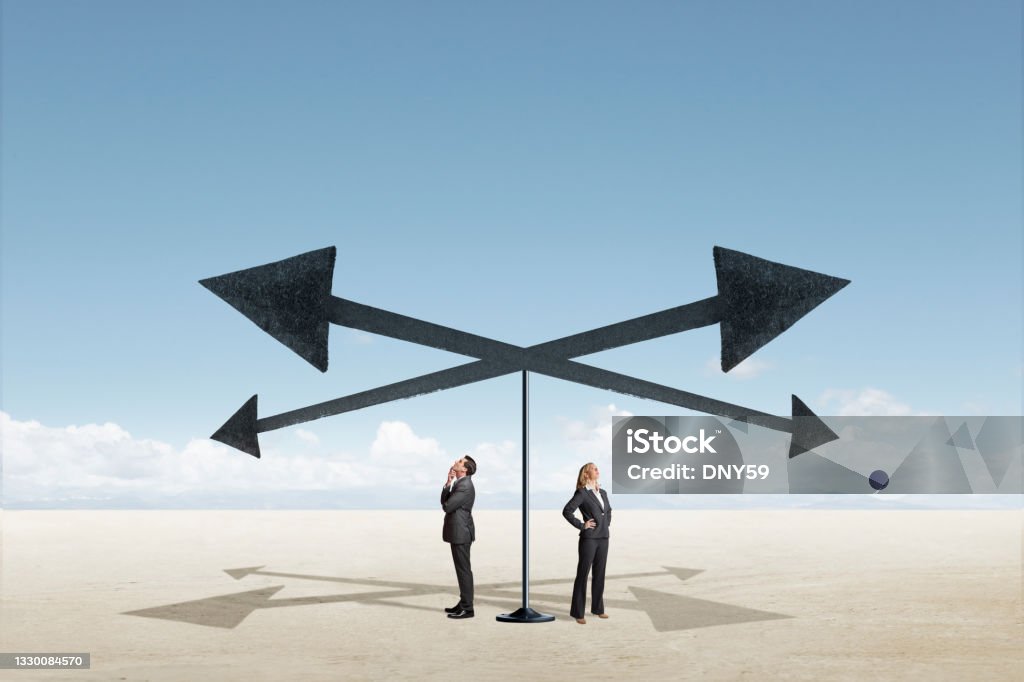 Man And Woman Stand At A Crossroad A man and a woman look up as they stand below four arrows pointing in different direction. Crossroads Sign Stock Photo