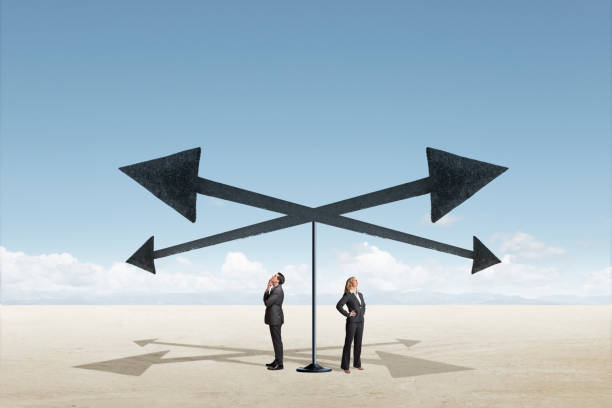 Man And Woman Stand At A Crossroad A man and a woman look up as they stand below four arrows pointing in different direction. crossroads sign stock pictures, royalty-free photos & images