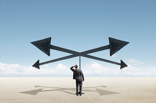 A man leans over to look out toward the horizon as he stands below four arrows pointing in different direction.