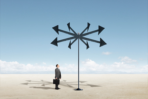 A man stands while holding his briefcase and looks up at a group of arrows on a pole that point in multiple directions.