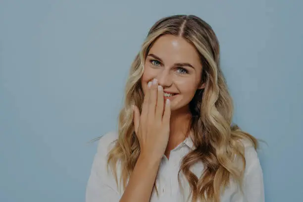 Attractive shy blonde woman being flattered while covering her mouth with hand, does not know what to say while standing isolated next to blue background. Human emotions concept