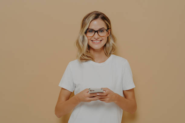 istock Young cute female student in spectacles using cell phone and looking at camera with pleasant smile 1330085000