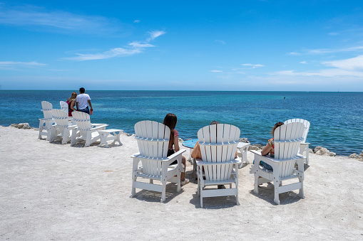 Islamorada, FloridaUSA - 06 13 2021. Outdoor sitting for beach dining at Lazy Days restaurant.  Family enjoying time together on vacation. People sitting on white wooden chairs with table on the beach with Atlantic ocean view.