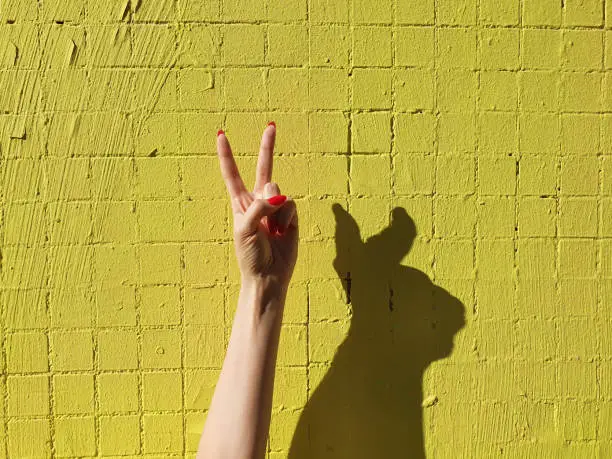 Photo of Peace sign hand. Woman's hand makes two fingers up gesture on neon green yellow background. Welcome gesture and rabbit shadow.