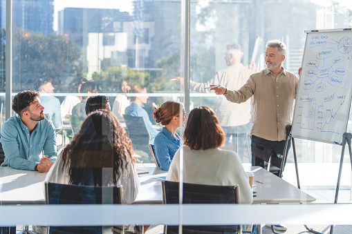 Business people watching a presentation on the whiteboard. A mature man is writing on the whiteboard with charts and graphs. They are sitting in a board room, there are laptop computers and technology on the table. All are casually dressed. There is a window behind him with city views.