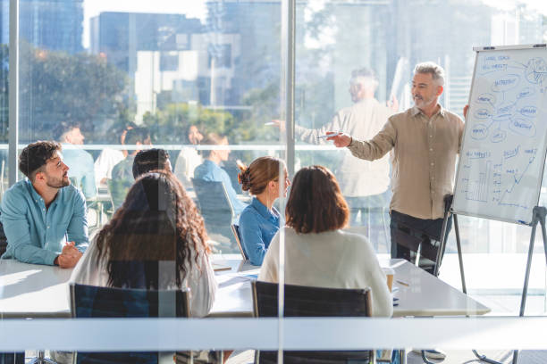 Business people watching a presentation on the whiteboard. Business people watching a presentation on the whiteboard. A mature man is writing on the whiteboard with charts and graphs. They are sitting in a board room, there are laptop computers and technology on the table. All are casually dressed. There is a window behind him with city views. training course stock pictures, royalty-free photos & images