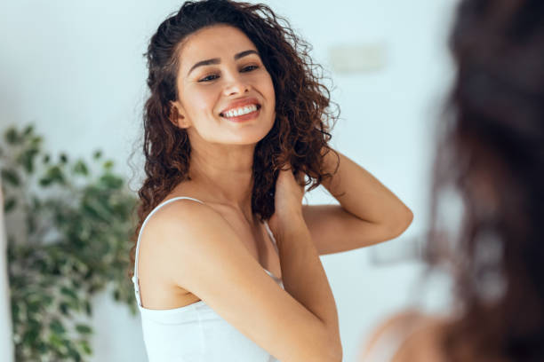 Beautiful young woman looking herself while smiling at mirror at home.