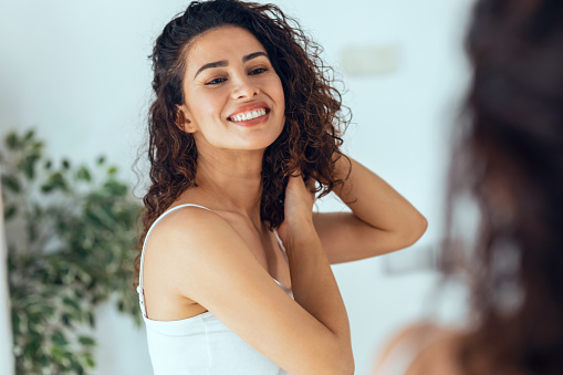 Portrait of beautiful young woman looking herself while smiling at mirror at home.