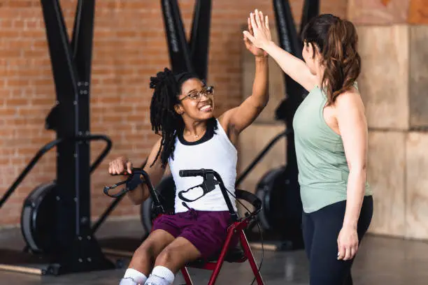 The young adult woman sitting in her wheelchair gives her mid adult female friend a high five after exercising together at the gym.