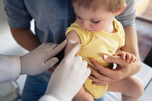 Unrecognizable doctor putting a patch on the little boy's shoulder after successful vaccination. Little boy sitting in father´s lap and looking at the patch.