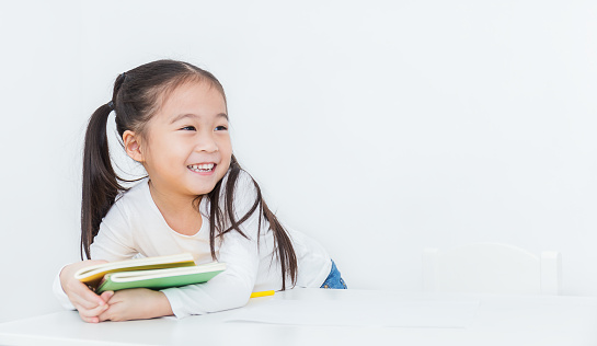 Close up portrait of little happy cute asian toddler girl sitting reading a book isolated on white background. Home school education study grade student back to school concept