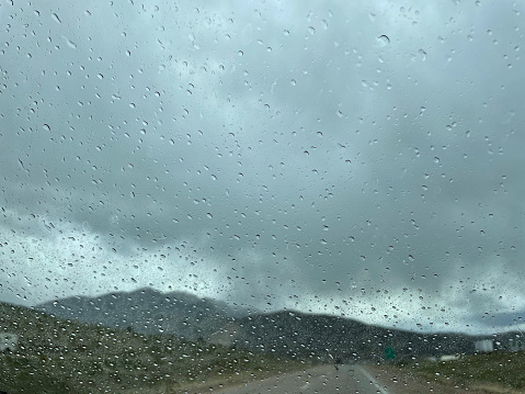 gray and blue clouds loom overhead and rain drops can be seen on windshield with mountains in the distance. Road ahead is out of focus. Focus is on the rain drops