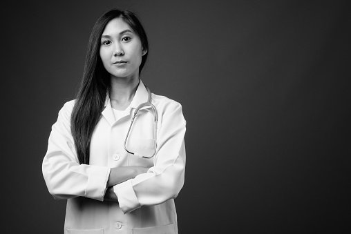 Studio shot of young beautiful Asian woman doctor against gray background in black and white
