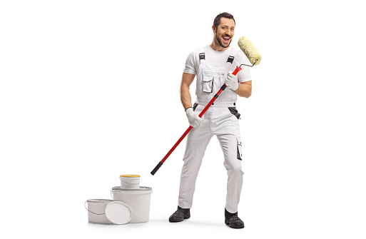 Full length portrait of a cheerful painter in a white uniform singing with a paint roller and a pile of buckets isolated on white background