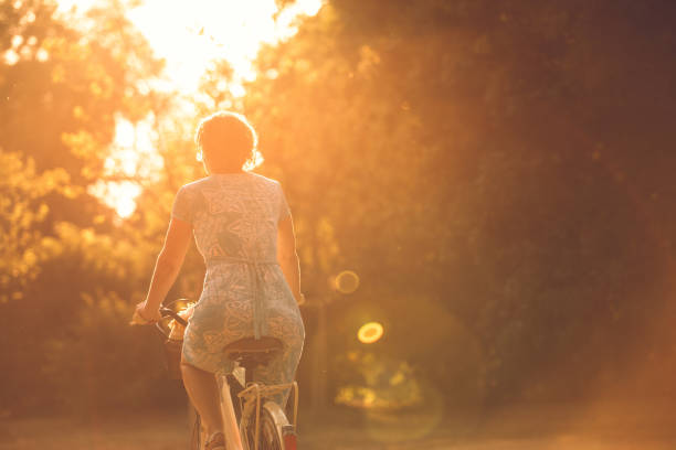 Riding her bike into the summer sunset Copy space shot of mature woman in a sundress sitting on her bike and riding into the sunset. golden hour stock pictures, royalty-free photos & images