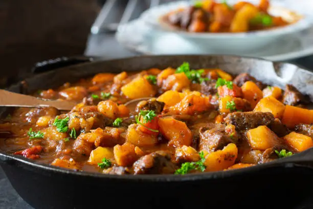 Traditional and fresh cooked irish stew with beef, carrots, potatoes and other vegetables served in a rustic cast iron pan on a table. Closeup view with blurred background