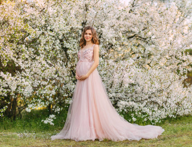 薄いピンクの長いイブニングドレスのポーズを着た若い美しい幸せな喜びの妊婦。咲く木々の背景。庭の緑は白い花を残します。女の子モデル。エレガンス妊娠春新鮮な自然 - weding gown ストックフォトと画像