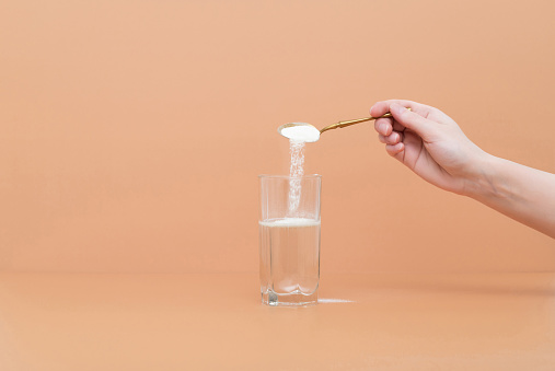 La mano vierte proteína de colágeno en polvo en un vaso de agua sobre un fondo beige. Un suplemento natural para la belleza de la piel y la salud ósea. photo