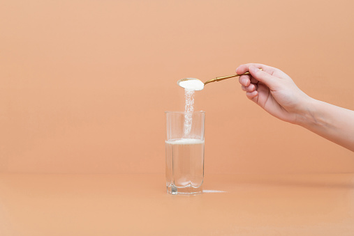 Hand pours collagen protein powder in a glass of water on a beige background. A natural supplement for skin beauty and bone health. Space for text.