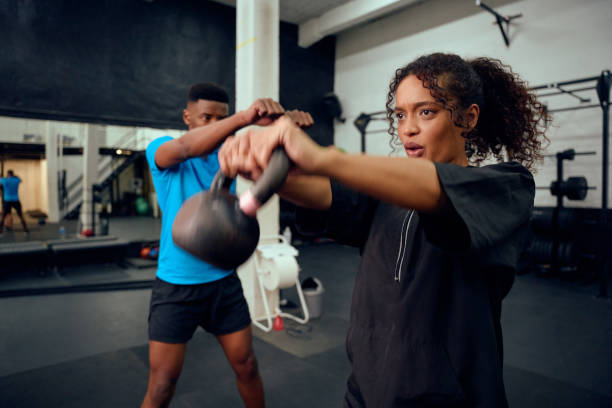 African American male personal trainer instructing African American female with a kettlebell routine in the gym. Mixed race friends doing cross training together. High quality photo African American male personal trainer instructing African American female with kettlebell exercise in the gym. Mixed race friends doing cross training together. High quality photo fitness trainer stock pictures, royalty-free photos & images
