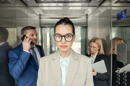 Portrait of young businesswoman in eyeglasses looking at camera while standing in elevator with other business people behind her back