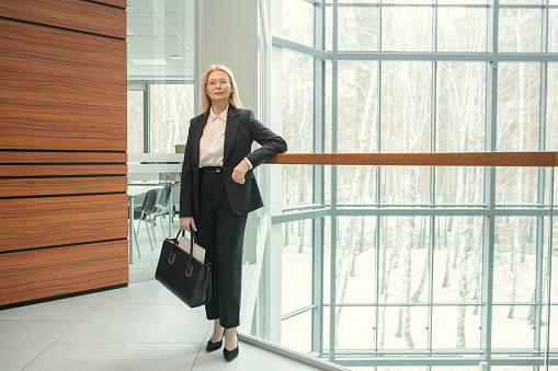 Portrait of confident mature businesswoman in elegant suit and with briefcase looking at camera while standing at modern office corridor