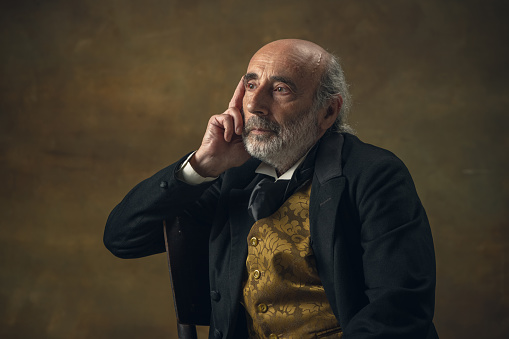 Thinking. Half-length retro portrait of elderly gray-haired man, gentleman or actor sitting on chair isolated on dark vintage background. Retro style, comparison of eras concept.