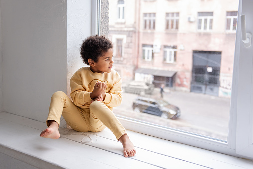A little African American boy in yellow pajamas sits barefoot on a wide windowsill by a large white window. The child is having fun, laughing and fooling around. Happy life style childhood at home.