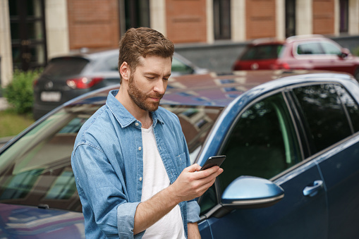 A handsome bearded man in a denim shirt and a white t-shirt is texting a message in the phone, leaning on his car, we see him from the waist up in a day light on a street