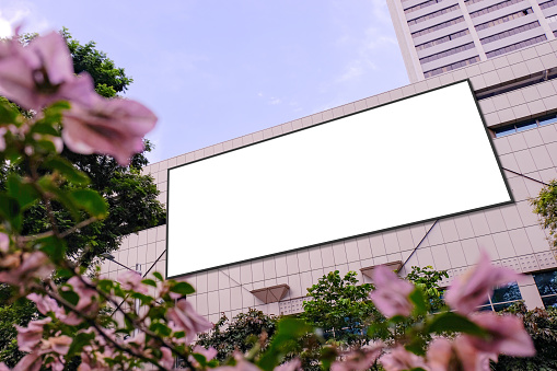 Blank advertising poster banner mockup on building exterior, flowers plants in foreground; large digital lightbox display screen. Billboard poster, out-of-home OOH media display space