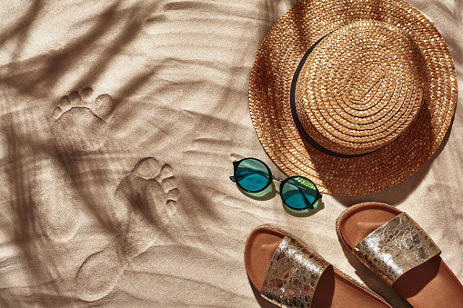 Summer concept with a shadow of a tropical palm tree leaves, copyspace. Traveler vacation accessories such as a straw hat, sunglasses and flip-flops are laid out on a white beach sand with foot print. Summertime lifestyle, objects in flat lay, top view arrangement.