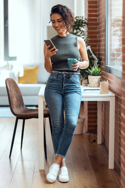 istock Smart business woman using mobile phone while drinking a cup of coffee leaning on the table in living room at home. 1330198952