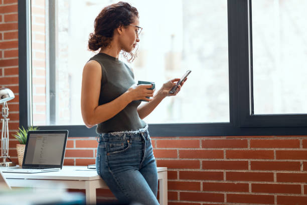 istock Smart business woman using mobile phone while drinking a cup of coffee leaning on the table in living room at home. 1330200029