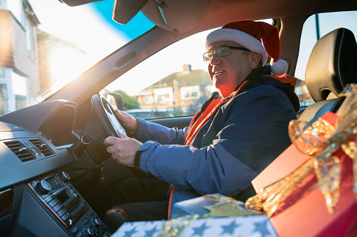 A medium close-up of a grandfather arriving at his grandkid's house with Christmas presents on the passenger seat in the car. He is wearing a Santa hat and some glasses. He has a content look on his face as he parks the car.