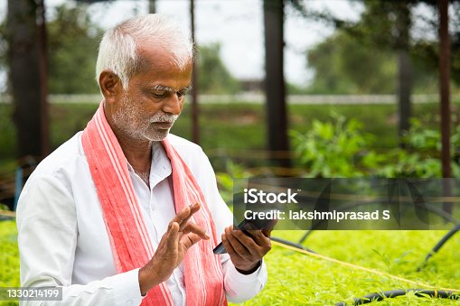 istock Indian Farmer busy using mobile phone while sitting in between the crop seedlings inside greenhouse or poly house - concept of farmer using technology and internet 1330214199