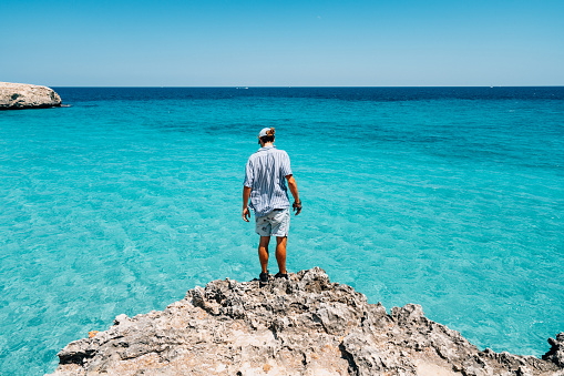 Young man on a cliff near a turquoise sea's water. He's standing, wearing a shirt.