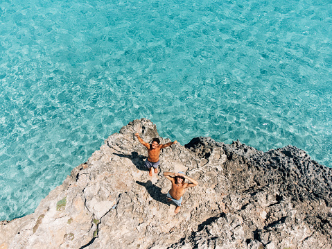 Two friends are looking up, saying hello to the drone. They are on a cliff near a turquoise sea.