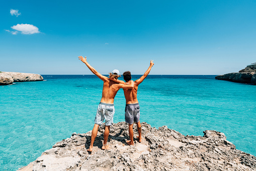 Two friends are standing on a cliff looking at the sea. They are embracing. Rear view. They are looking at view with open arms.