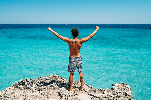 Rear view of a young adult man standing on a cliff with open arms. He's looking at view. Turquoise sea's water in the background.