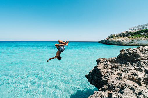 Young man is diving in the sea from a cliff doing a backflip. Carefree, vacation, summer, freedom concept.
