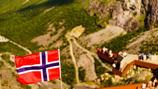 Trollstigen mountain road landscape in Norway, Europe. Norwegian flag waving and many tourists people on viewing platform in background. National tourist route.