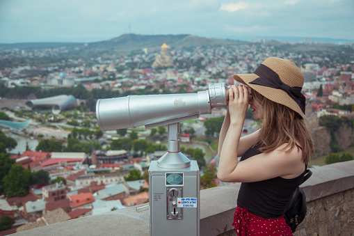 Woman with hat looking through binoculars to Tbilisi city